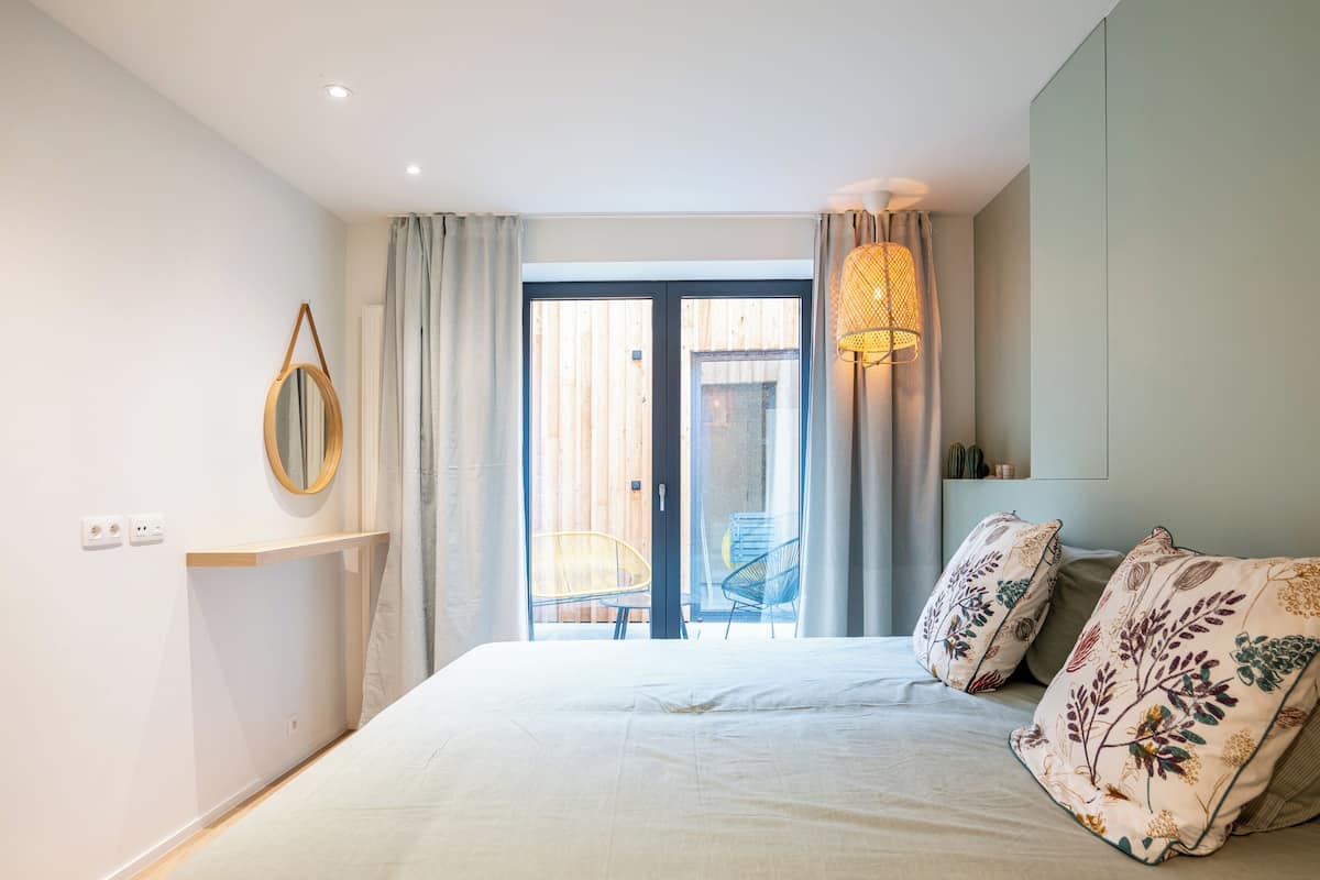 A bedroom with a bed, a bedside table, a mirror, a window with curtains, and a view of the outside.