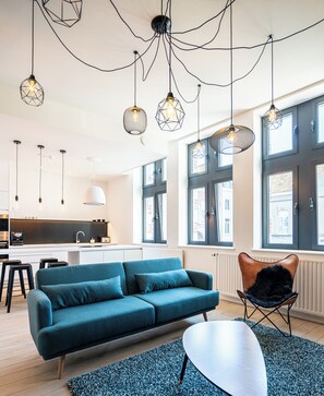 Ein modernes Wohnzimmer mit einem türkisfarbenen Sofa, einem schwarzen Ledersessel und einem weißen Couchtisch. Von der Decke hängen Pendelleuchten mit geometrischen Mustern.