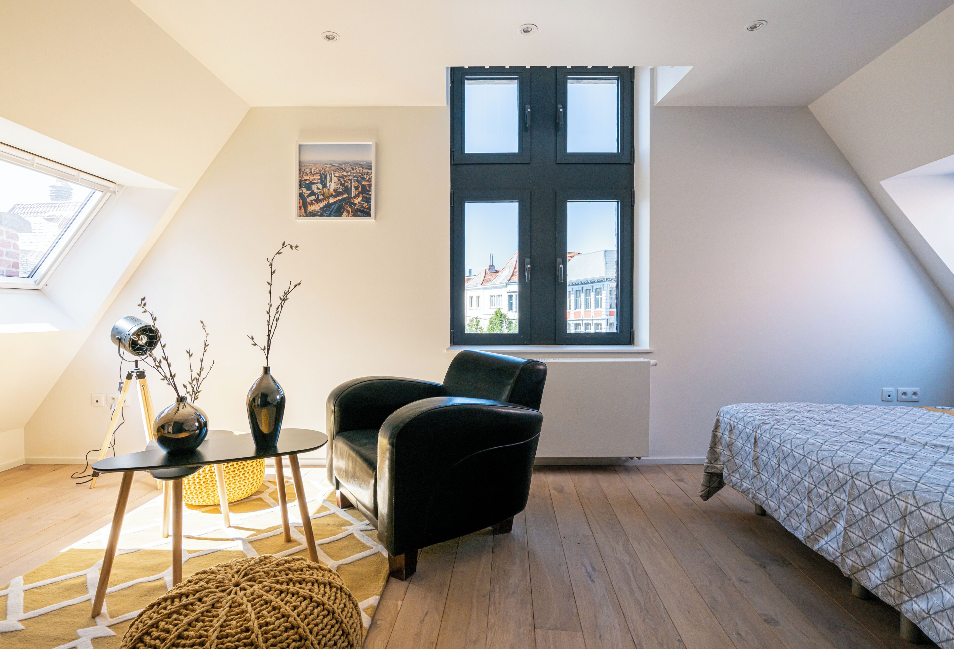 A modern loft bedroom with a large window, a black leather chair, a bed with a patterned duvet, and a cityscape view.