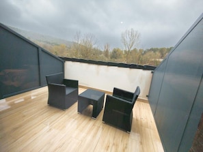 A rooftop terrace with two black wicker chairs and a small table, wooden flooring, and a view of trees and hills under a cloudy sky.