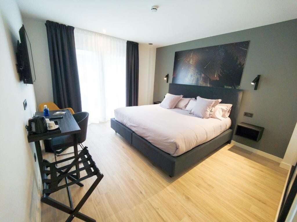 Ett modernt hotellrum med en säng, ett skrivbord och en stol, ett stort fönster med gardiner och en tavla på väggen.