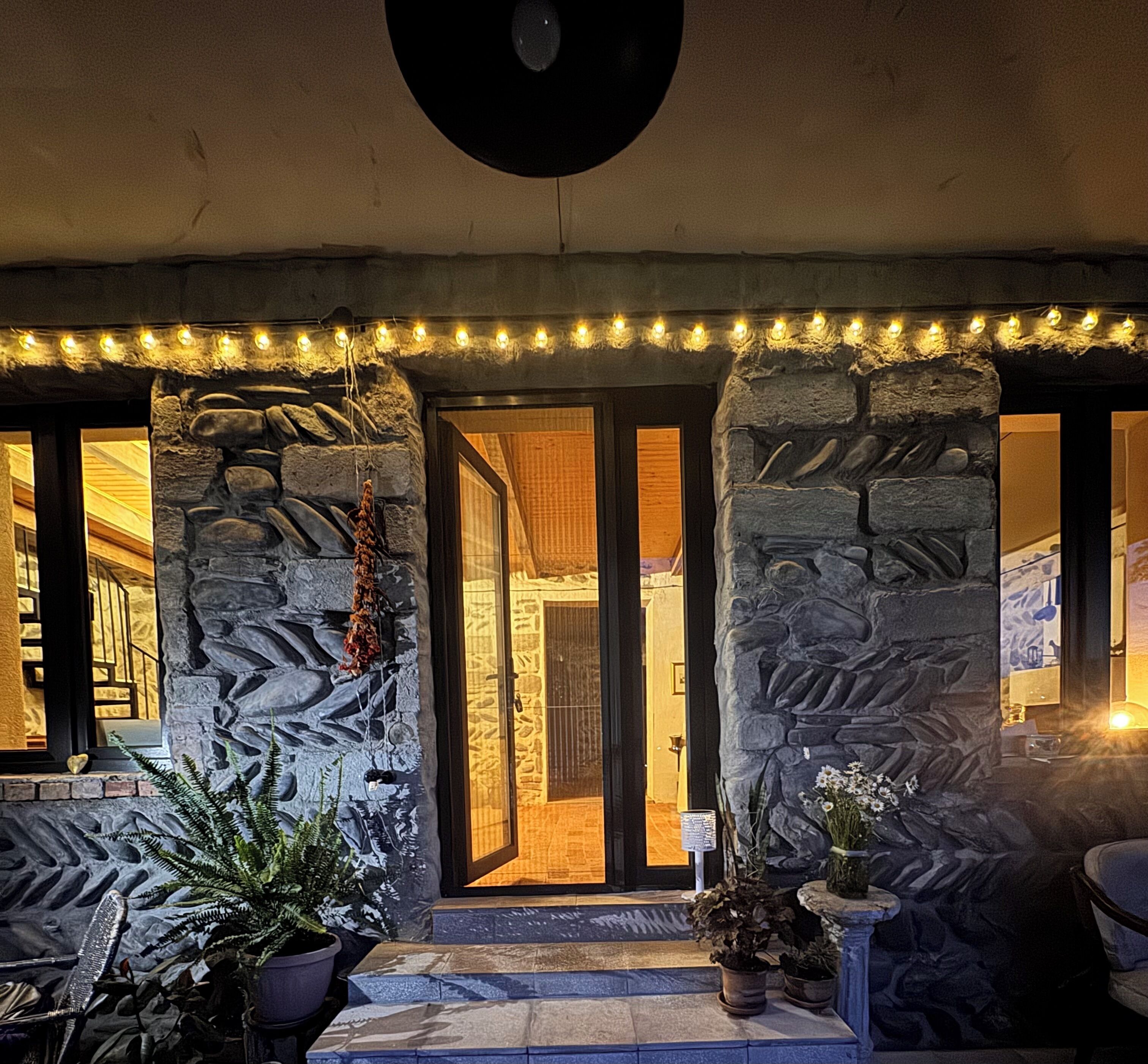 Une façade en pierre avec une porte d’entrée, des plantes en pot et des guirlandes lumineuses.