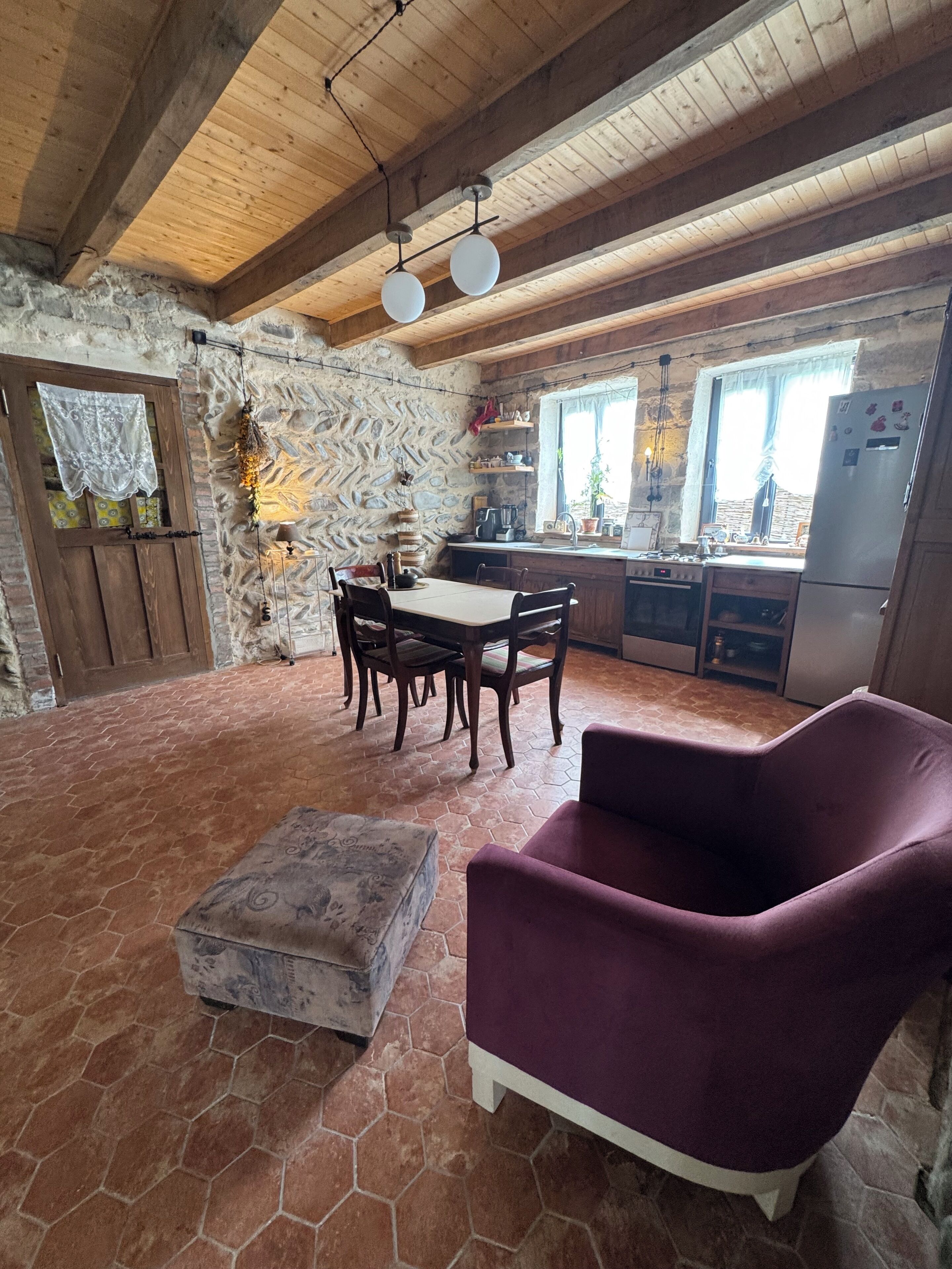 Une cuisine rustique avec un mur en pierre, des poutres en bois, une table à manger avec des chaises, un fauteuil violet et un sol carrelé.