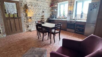 Une cuisine rustique avec un mur en pierre, des poutres en bois, une table à manger avec des chaises, un fauteuil violet et un sol carrelé.