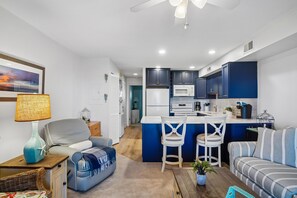 Eine moderne Küche mit dunkelblauen Schränken, einem weißen Kühlschrank und einer Mikrowelle. Es gibt einen Essbereich mit einem weißen Stuhl und einem blauen Sofa.