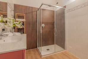 Une salle de bain avec une douche à parois de verre, un lavabo blanc et une étagère en bois où sont posées des serviettes.