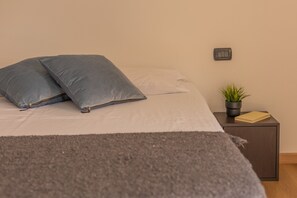 Ein ordentlich bezogenes Bett mit Kissen, ein Nachttisch mit Pflanze und eine Steckdose an der Wand.