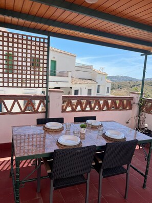 Terraço com mesa posta para refeições, cadeiras de vime e vista para edifícios ao fundo.