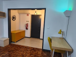 Una habitación con suelo de madera, un escritorio con lámpara, un banco, un extintor y una puerta.