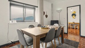 A dining area with a wooden table, grey chairs, a window with curtains, a TV, and a wall-mounted light fixture. - Cantinho do Préstimo (Préstimo)