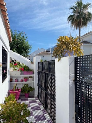 A narrow outdoor corridor with potted plants, a tiled floor, and a black metal fence.