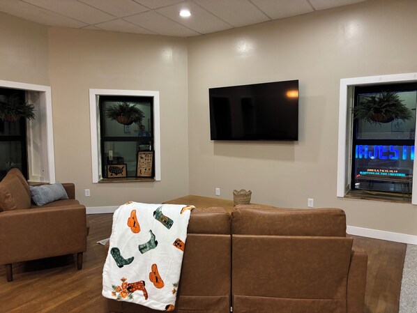 Una sala de estar con un sofá de cuero marrón, un televisor de pantalla plana montado en la pared y ventanas con plantas decorativas.