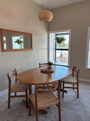 Comedor con una mesa redonda de madera, cuatro sillas de mimbre, una lámpara colgante, un espejo y un ventanal con vistas a edificios.