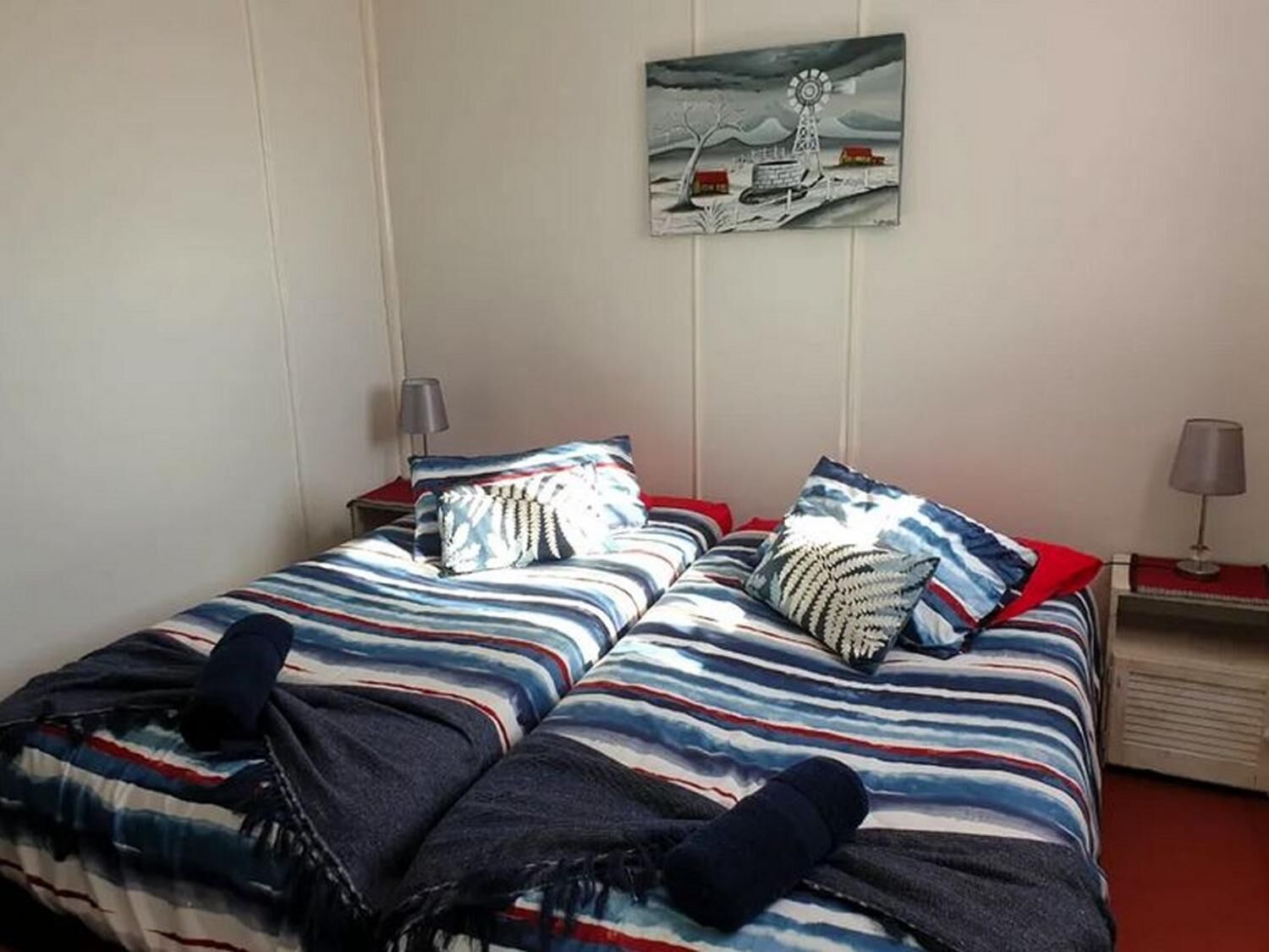 Een tweepersoonsbed met gestreepte beddengoed, twee nachtkastjes met lampen, en een aan de muur hangend schilderij van een windmolen.