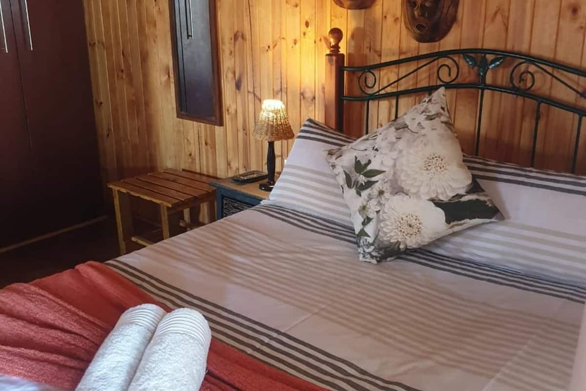 Una habitación con paneles de madera, una cama, una mesita de noche, una lámpara y un taburete de madera pequeño.