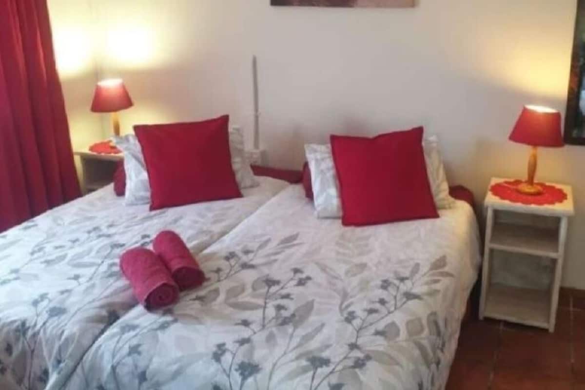 Cama doble con ropa de cama floral y almohadas rojas, dos mesitas de noche con lámparas y una cortina roja.