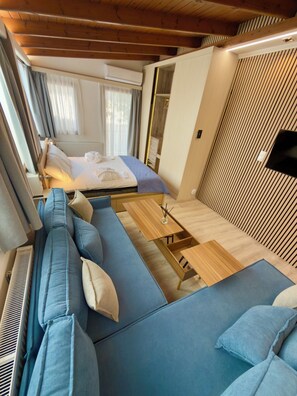 A cozy RV interior with a bed, blue sofa, wooden tables, and a TV mounted on the wall. - Apartment Vagia Nea Smyrni (Nea Smirni)