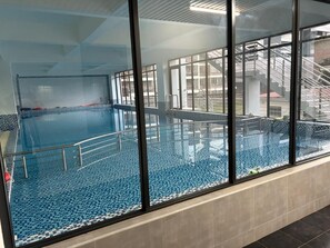 Una piscina interior con paredes de vidrio transparente, suelo de baldosas y baranda de metal.