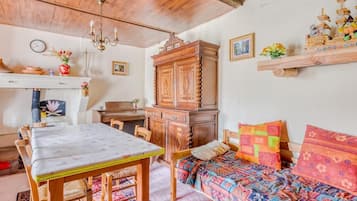 Une pièce chaleureuse avec un plafond en bois, une table à manger, un lit avec une literie colorée, une armoire en bois et une étagère ornée d’objets décoratifs.