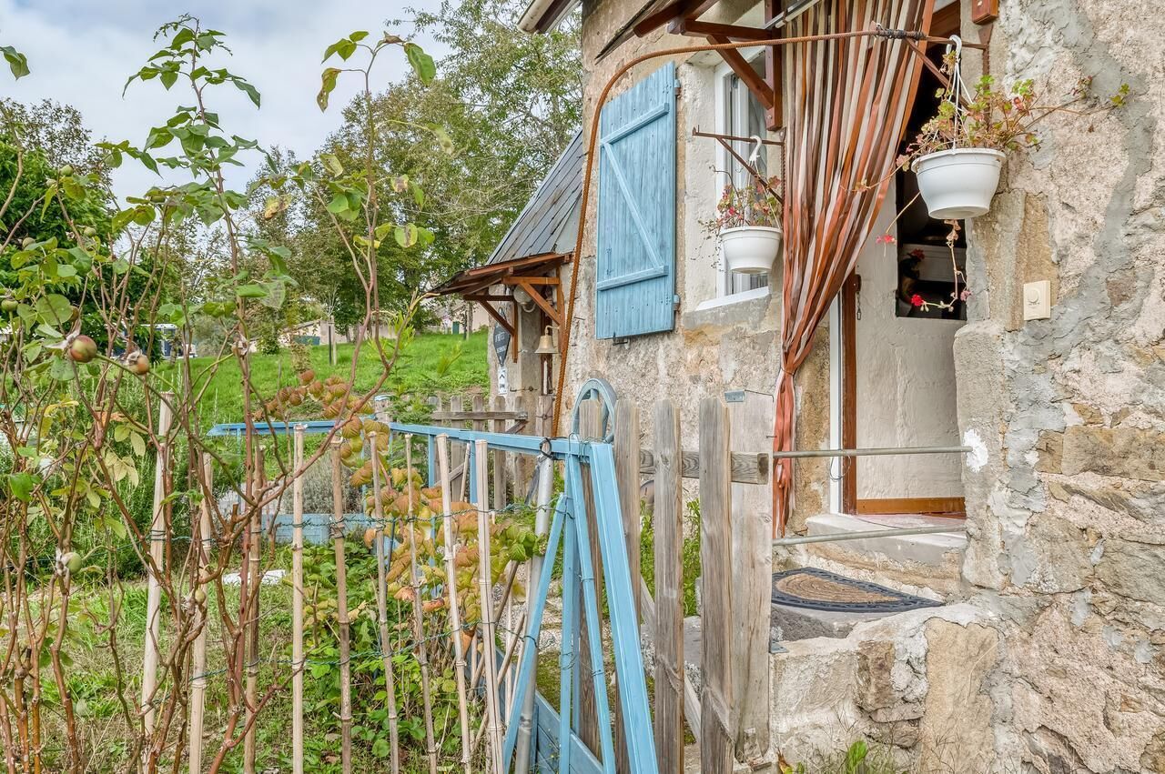 Une maison de campagne en pierre, dotée d’un porche en bois, d’un volet bleu et d’un pot de fleurs suspendu.