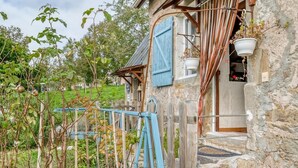 A rustic stone house with a wooden porch, a blue shutter, and a hanging planter.
