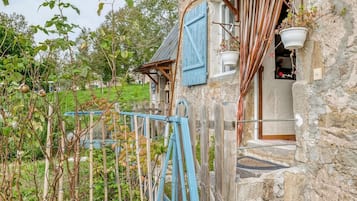 Une maison de campagne en pierre, dotée d’un porche en bois, d’un volet bleu et d’un pot de fleurs suspendu.
