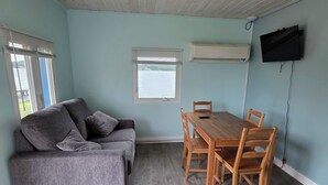 A room with a grey sofa, wooden dining table with chairs, a window with blinds, an air conditioning unit, and a flat-screen TV mounted on the wall. - Cozy 2-bedroom cottage in marvelous Ludington with AC (Ludington)