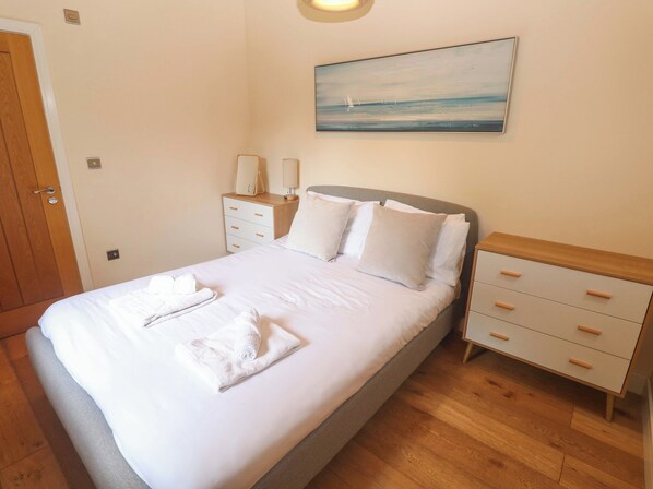 Een opgemaakt bed met witte lakens en handdoeken, een nachtkastje met een lamp, een wandlamp en een schilderij van een zeegezicht aan de muur.