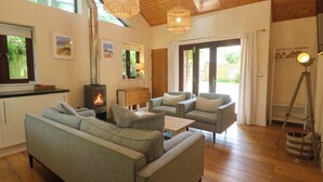 A cozy living room with a sofa, armchairs, a wooden floor, a fireplace, and a large window. - The Lodge at Lamburn (Truro)