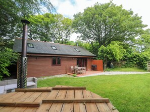 Eine Holzterrasse mit Grill und Gartenmöbeln, ein Haus mit Satteldach und einem gepflegten Rasen.
