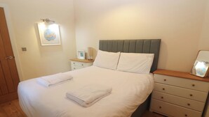 A neatly made bed with white linens, a bedside table with a lamp, and a small bedside table with a picture frame. - The Lodge at Lamburn (Truro)