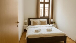 Ein ordentlich bezogenes Bett mit weißen Leinentüchern und zusammengerollten Handtüchern, ein Holzboden, ein Fenster mit Vorhängen und ein Nachttisch mit Lampe.