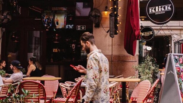 Ein Straßencafé mit Korbstuhlen und -tischen. Ein Mann geht vorbei, im Hintergrund ein Schild für 'KIKK BISTROBAR'.