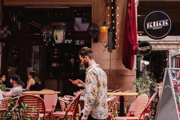 An outdoor cafe with wicker chairs and tables, a man walking by, and a sign for 'KIKK BISTROBAR' in the background.