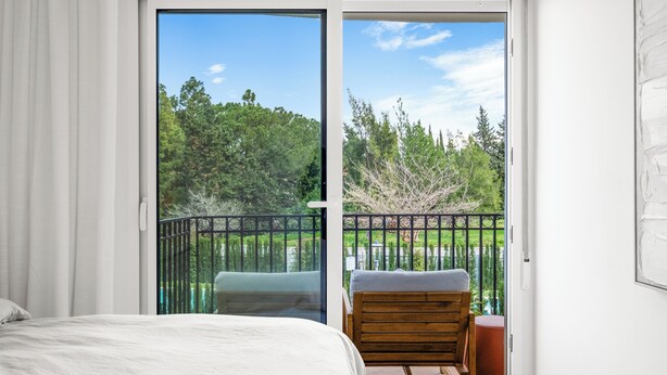 Una camera da letto con un letto grande, una sedia di legno e un balcone affacciato su alberi e un recinto.