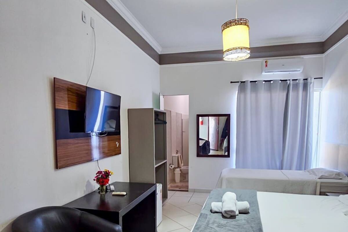 A hotel room with a bed, a desk, a chair, a TV, a mirror, and a hanging light fixture.
