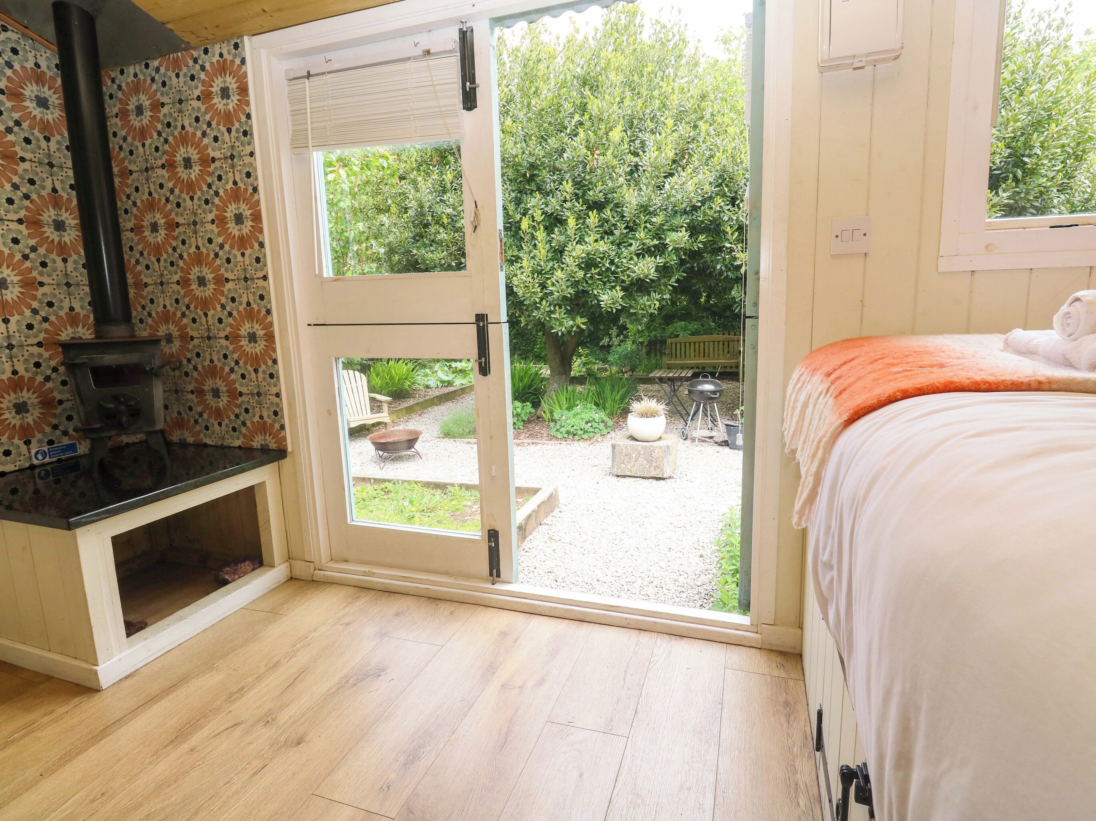 A room with a wooden floor, a bed with an orange blanket, a white door leading to a garden, and a fireplace.