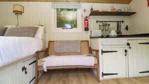 A room with a bed, a wooden bench, a window with blinds, a shelf with kitchen items, and a fire extinguisher. - Shepherds Hut (Truro)