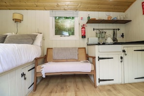 Ein Zimmer mit einem Bett, einer hölzernen Bank, einem Fenster mit Jalousien, einem Regal mit Küchenutensilien und einem Feuerlöscher.