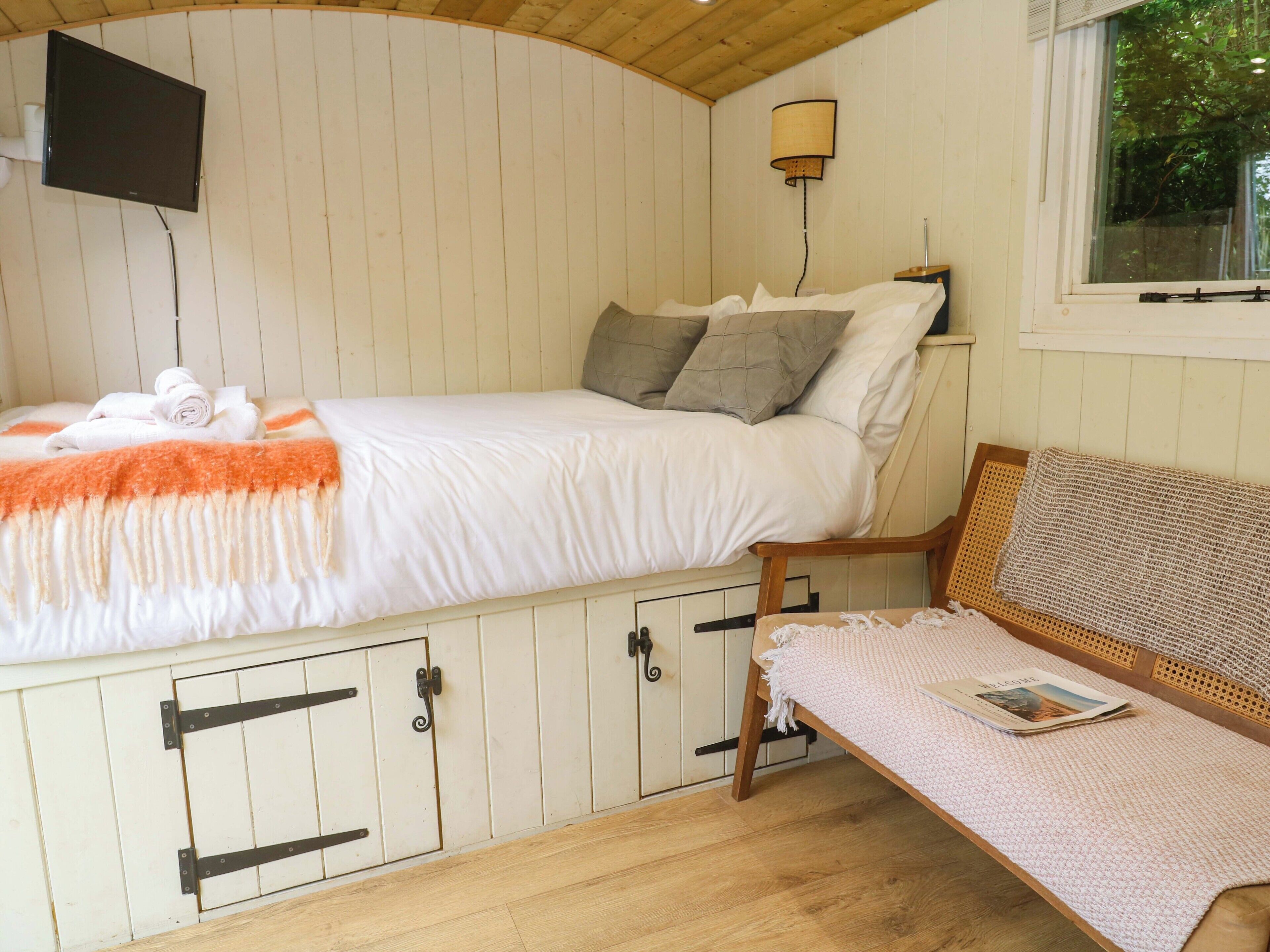 A single bed with a white mattress and orange blanket, a wooden bench with a magazine, a wall-mounted TV, and a window with a view of greenery.