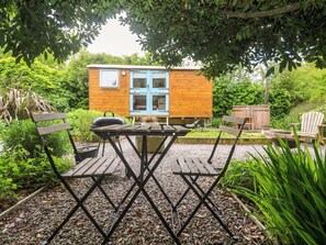 Eine Holzhütte mit blauer Tür und Fenstern, umgeben von Grünpflanzen und einer gepflasterten Terrasse mit Tisch und Stühlen.