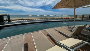 Ein Poolbereich mit Liegestühlen und Sonnenschirmen, Blick auf einen Strand und ein klarer Himmel.