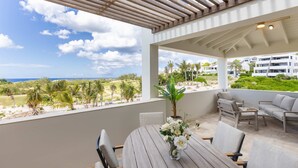 Un espace extérieur abrité, équipé d’une table et de chaises, d’une plante en pot, et offrant une vue sur la plage et les bâtiments.