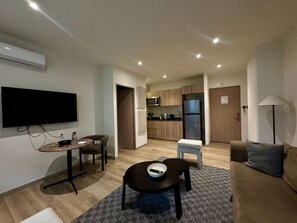 Ein modernes Wohnzimmer mit Flachbildfernseher, Esstisch mit Stühlen, einer Couch sowie einer Küchenzeile inklusive Kühlschrank und Mikrowelle und einer Lampe.
