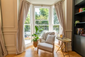 Ein Zimmer mit Erkerfenster, einem Sessel, einem kleinen Tisch und einem Bücherregal.