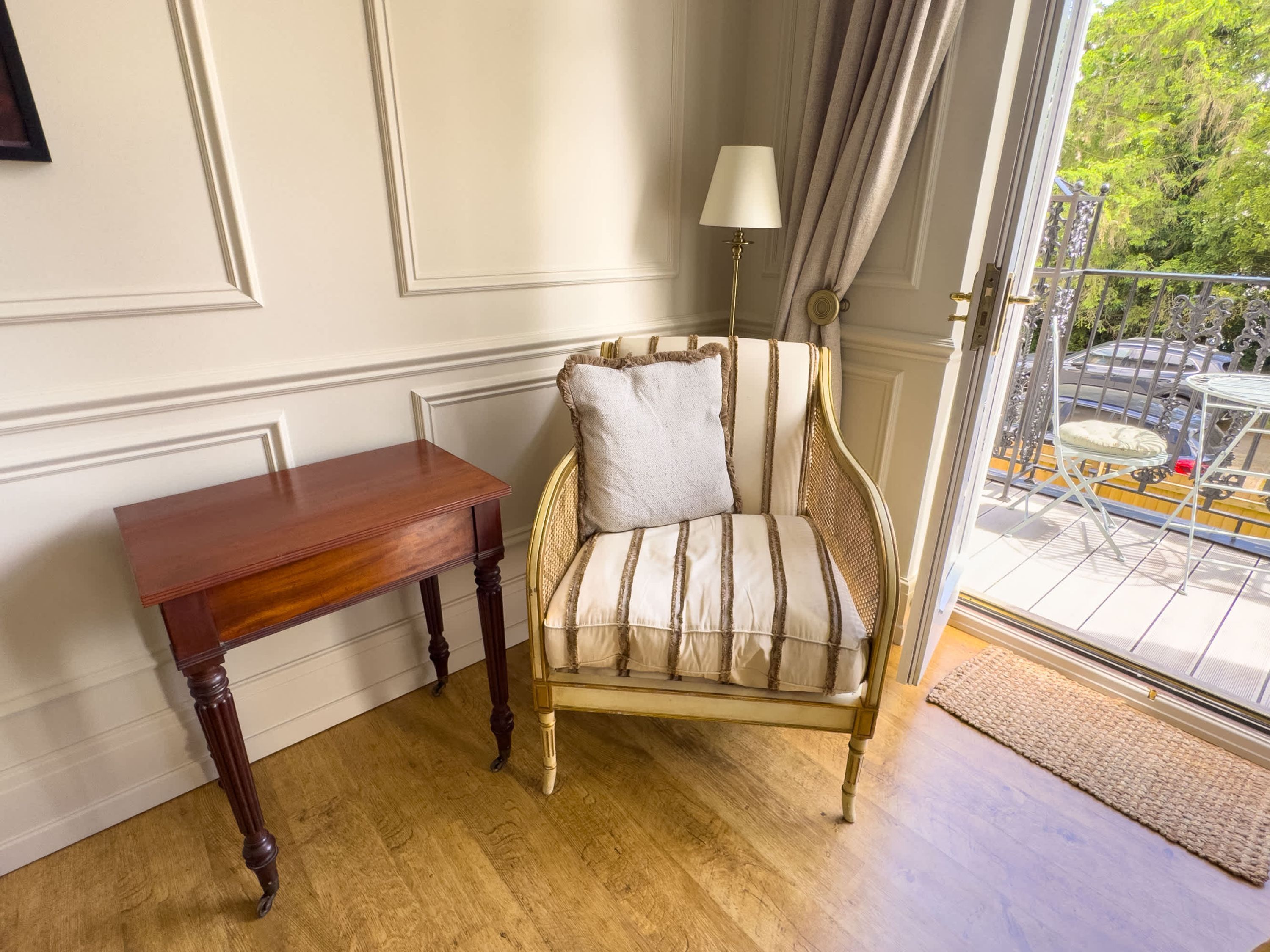 A corner room with a wooden table, a striped armchair, a floor lamp, and a view of a balcony with outdoor furniture.