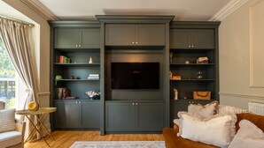 Uma sala de estar com um painel de entretenimento verde escuro, uma TV de tela plana, um sofá marrom e uma mesa redonda pequena com um abajur.