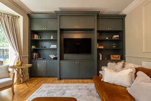 Ein Wohnzimmer mit einer dunkelgrünen Unterhaltungsecke, einem Flachbildfernseher, einem braunen Sofa und einem kleinen runden Tisch mit einer Lampe.