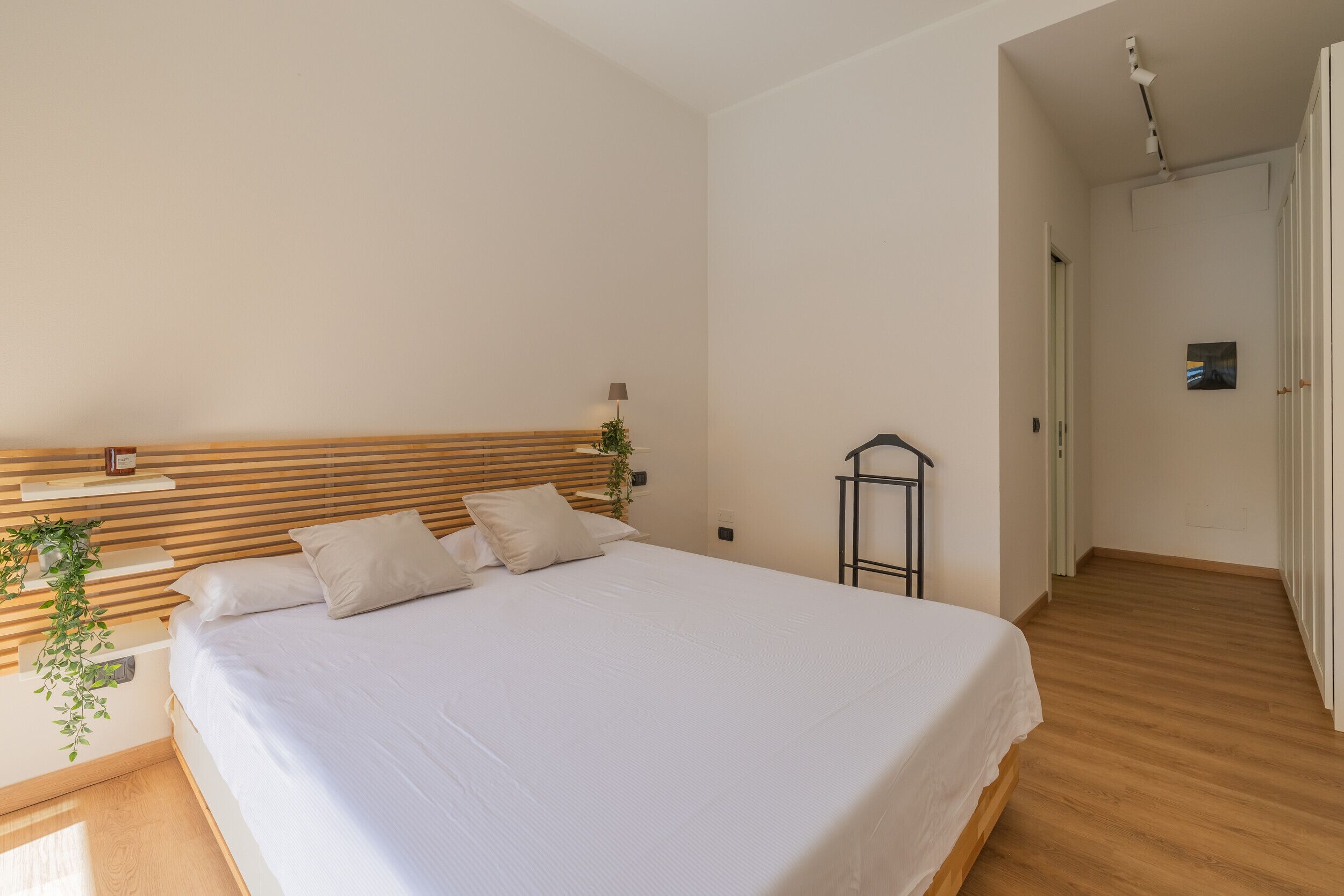 Un dormitorio con cabecera de madera, cama con ropa de cama blanca, mesita de noche con lámpara y una planta.