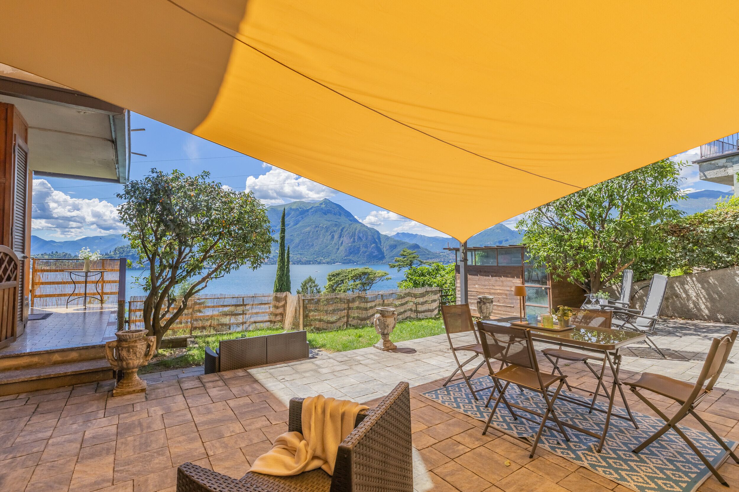 Un patio con tavolo e sedie, un divano in vimini, un baldacchino giallo e vista su un lago e montagne.